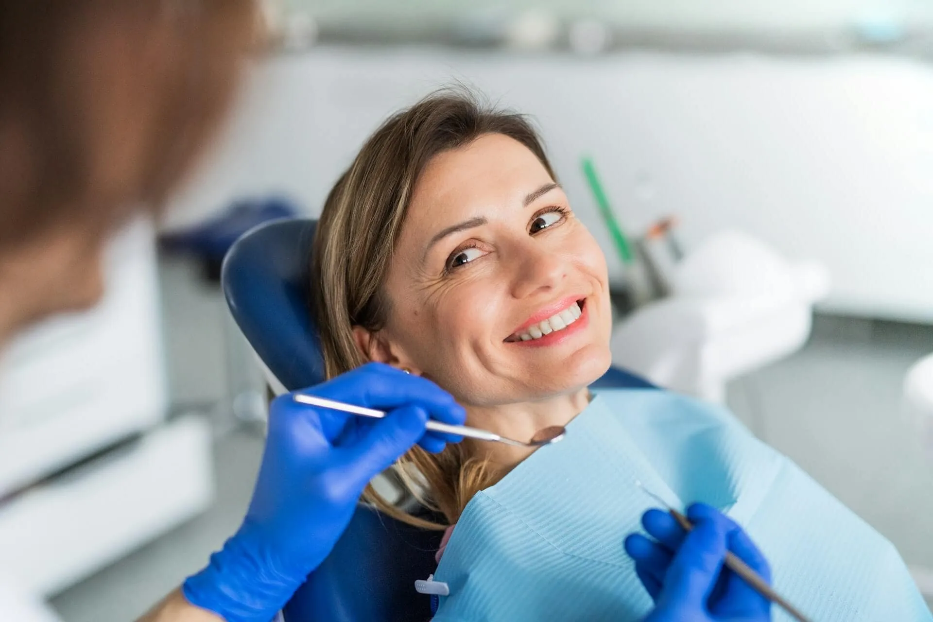 Smiling dental patient in chair during exam with dentist wearing blue gloves and holding dental tools.
